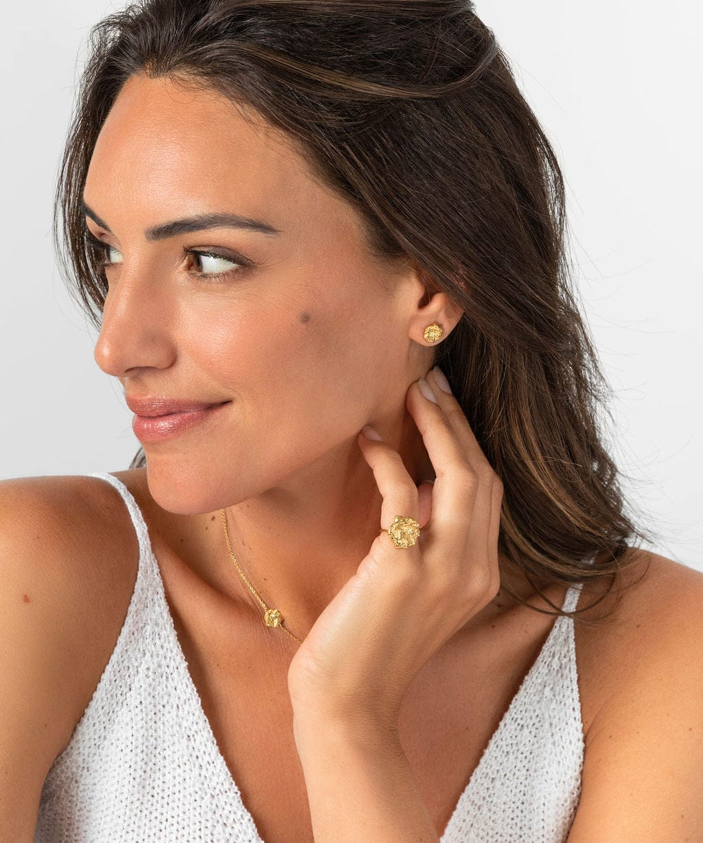 A woman wearing a gold-toned stud earring and a matching textured, circular ring on her finger while touching her neck.